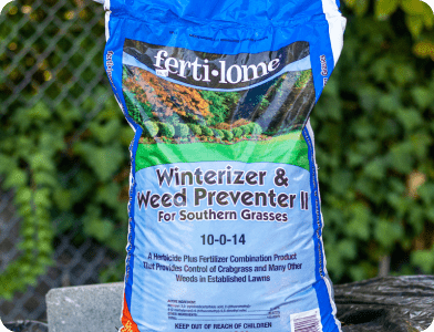 A blue and white bag of ferti-lome Winterizer & Weed Preventer II for Southern Grasses sits outdoors. The bag is labeled 10-0-14 and is designed for preventing weeds and fertilizing lawns.