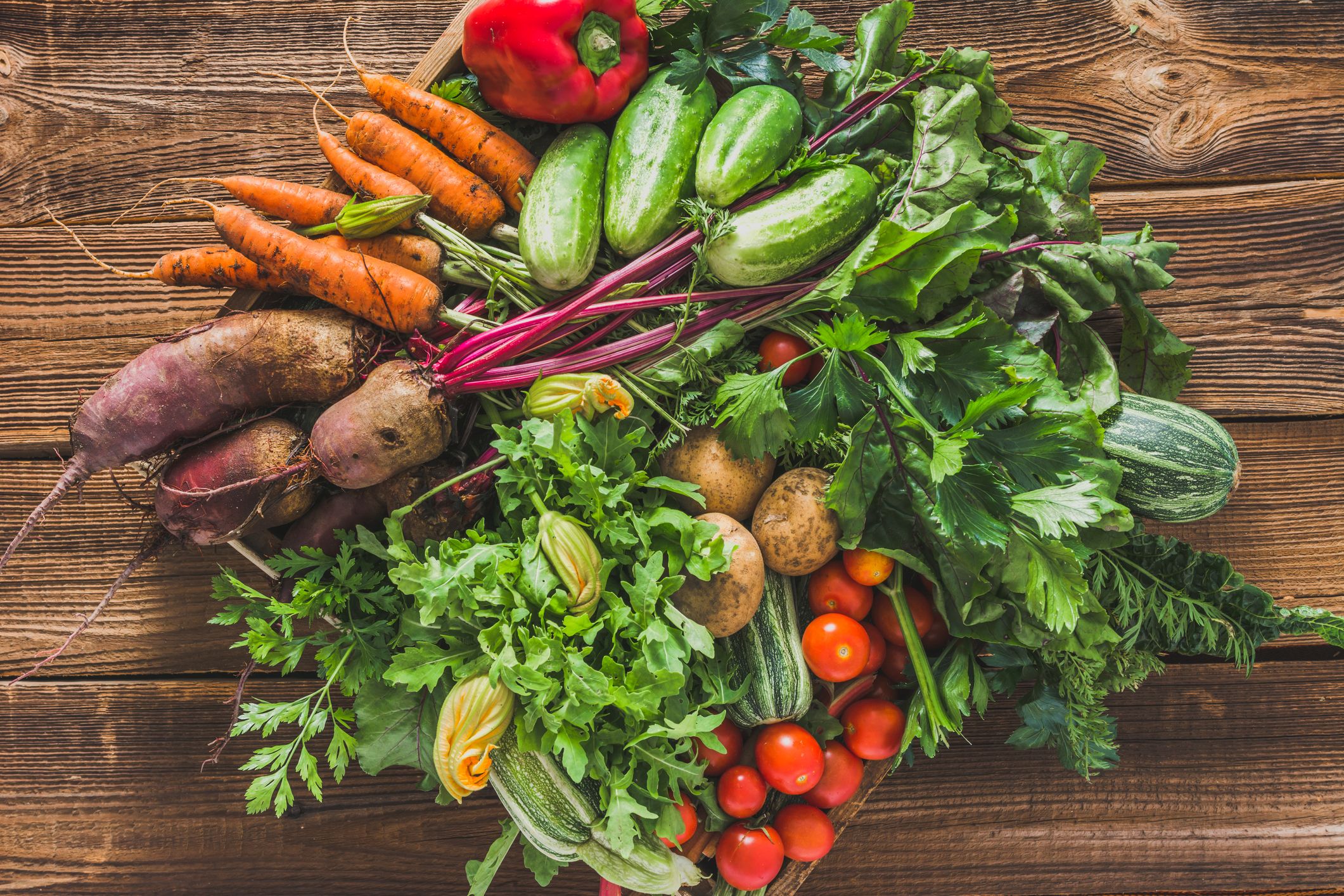 gettyimages-1030722720 An assortment of fresh vegetables, including carrots, cucumbers, tomatoes, leafy greens, potatoes, beets, and a red bell pepper, arranged on a rustic wooden surface.