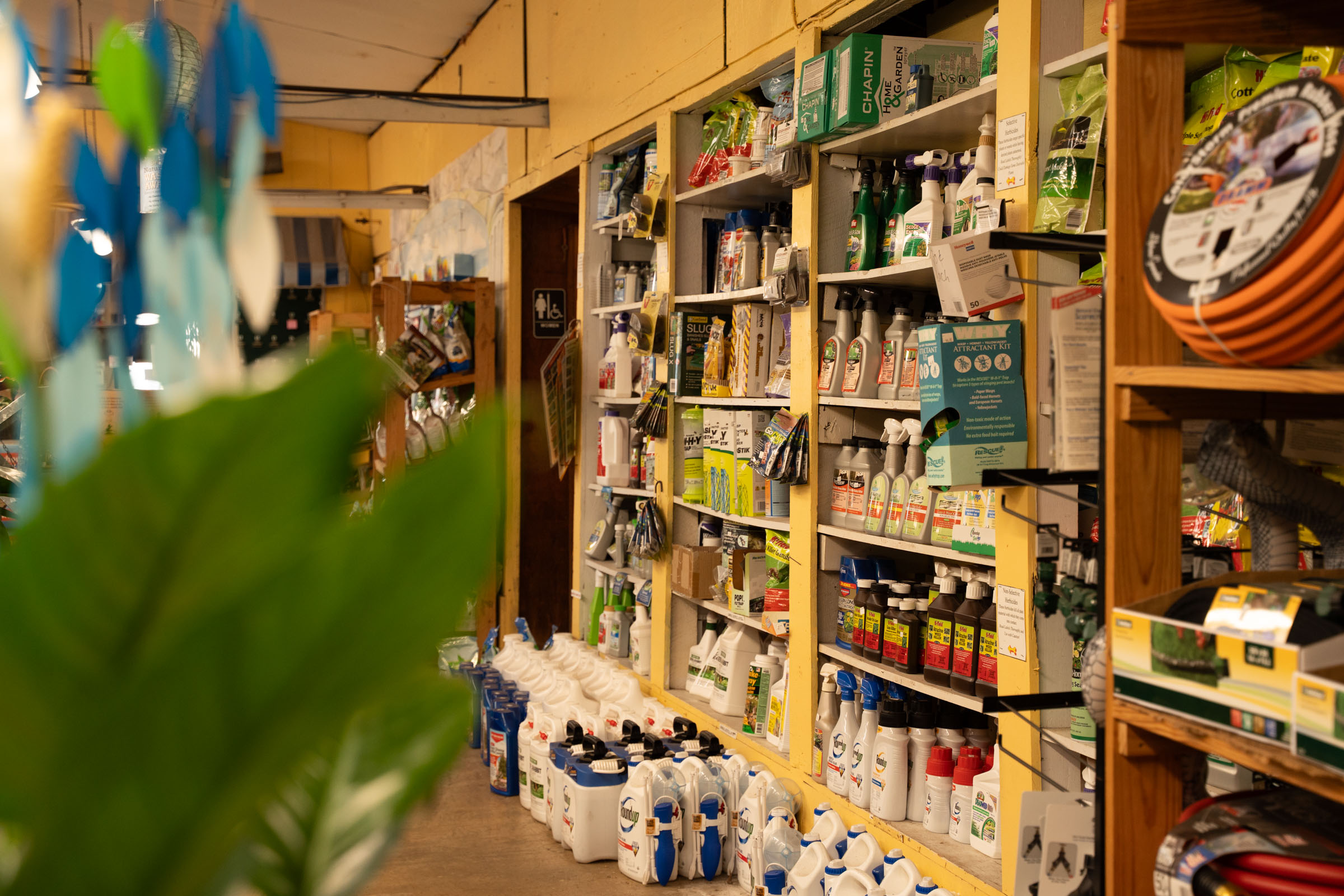 A store aisle with shelves stocked with gardening supplies, including bottles, sprays, and containers. Large white jugs and various products line the bottom shelf. A leafy green plant is partially visible in the foreground.